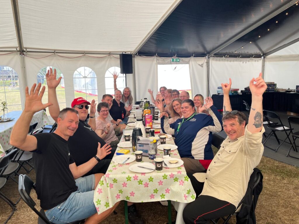Deltagere vinker fra et langt bord under et måltidsfællesskab i et telt